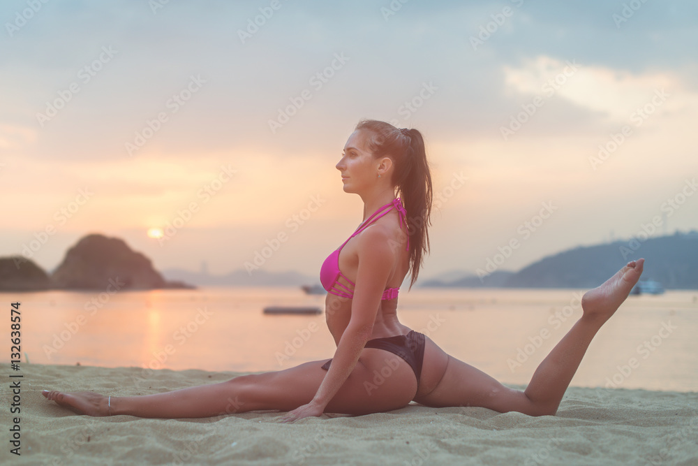 Photo in profile of slim fitness model with brown hair wearing bikini doing leg split exercise on beach at sunrise against sea, sky and mountains
