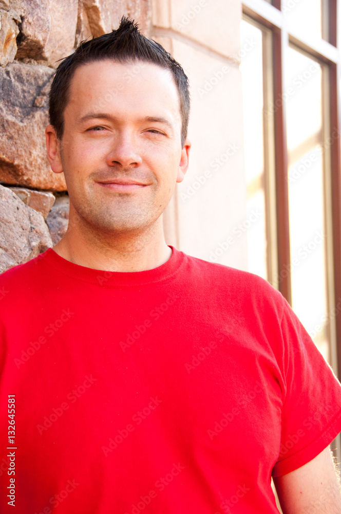 Obraz premium Young man leans against a building with a rock wall and smiles at the camera. Man wears a red tee shirt, dark hair, caucasian man in this late 20s.