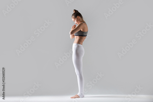 Young attractive woman practicing yoga, standing in mountain exercise, Tadasana pose, namaste, working out wearing sportswear, white pants, top, indoor full length, isolated, grey studio background