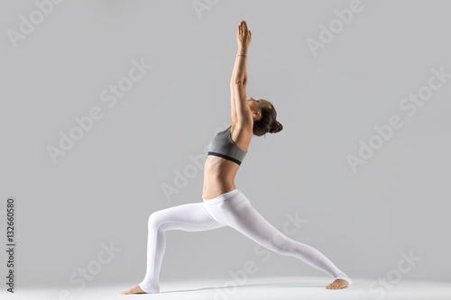 Young attractive woman practicing yoga, standing in Warrior one exercise, Virabhadrasana I pose, working out wearing sportswear, indoor full length, isolated against grey studio background