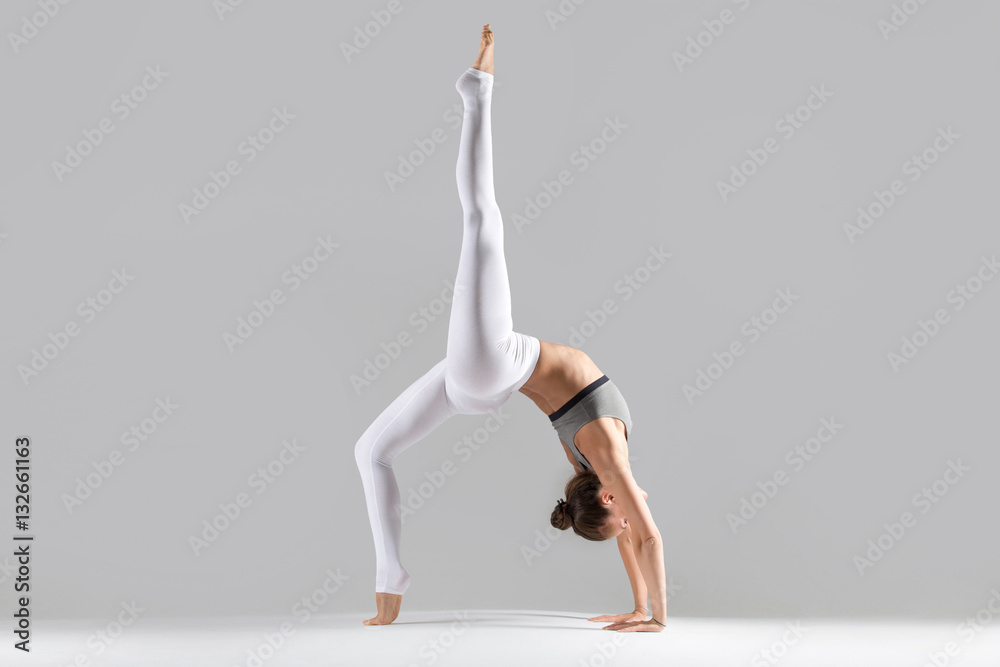 Young attractive woman practicing yoga, standing in Bridge exercise, One legged Wheel pose, working out wearing sportswear, white pants, indoor full length, isolated against grey studio background