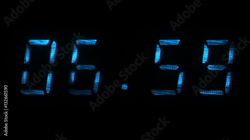 Digital clock with fluorescent display shows the time of 06 hours 59 minutes to 07 hours 00 minutes in the blue on a black background photo