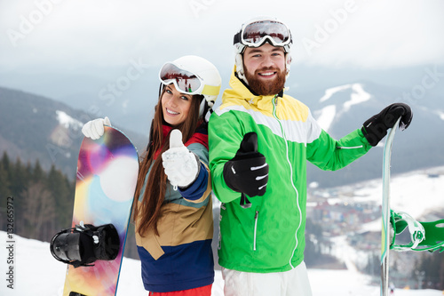 Cheerful loving couple snowboarders on slopes make thumbs up gesture