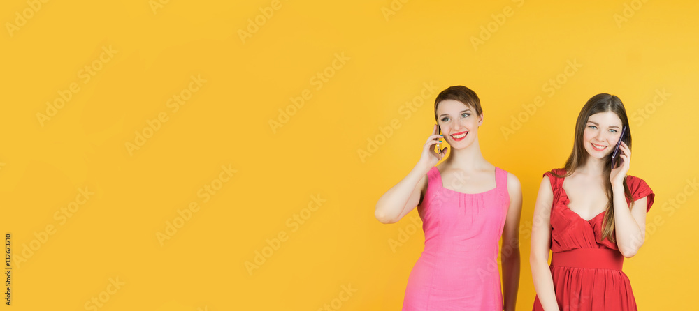 Technology, friendship and leirure concept - two smiling young women talking with smartphones. Isolated studio, yellow background, female model. Banner. Technology, friendship and leirure concept - two smiling young women talking with smartphones. Isolated studio, yellow background, female model. Banner.