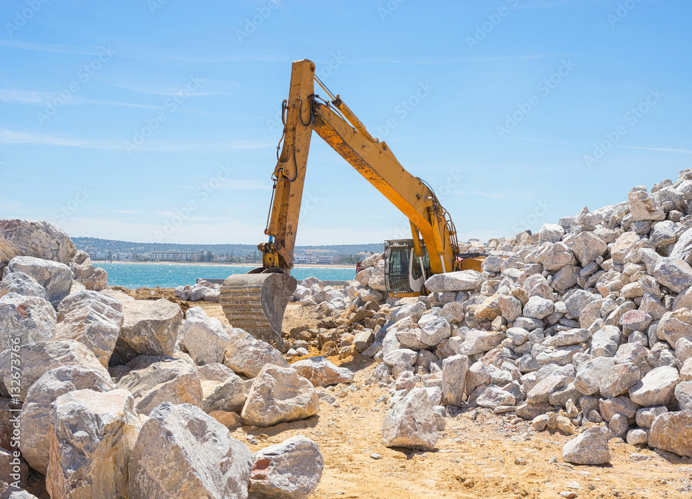Mechanical excavator working on coast with stones.