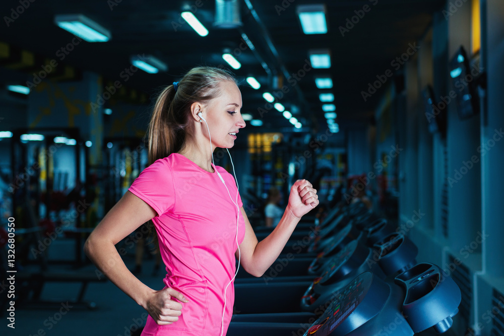 Obraz premium Attractive fitness girl running on machine treadmill. Pretty girl doing workout at modern fitness gym.