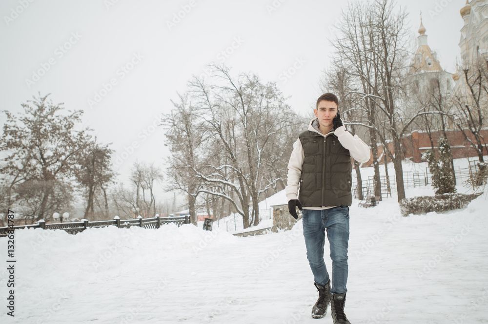 Outdoor winter portrait for young handsome man with the phone. Beautiful teenager in his jacket and vest posing on a city street, background of fir trees.