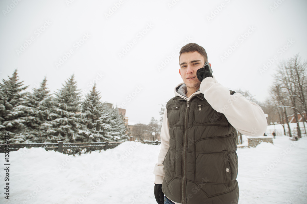 Outdoor winter portrait for young handsome man with the phone. Beautiful teenager in his jacket and vest posing on a city street, background of fir trees. Outdoor winter portrait for young handsome man with the phone. Beautiful teenager in his jacket and vest posing on a city street, background of fir trees.