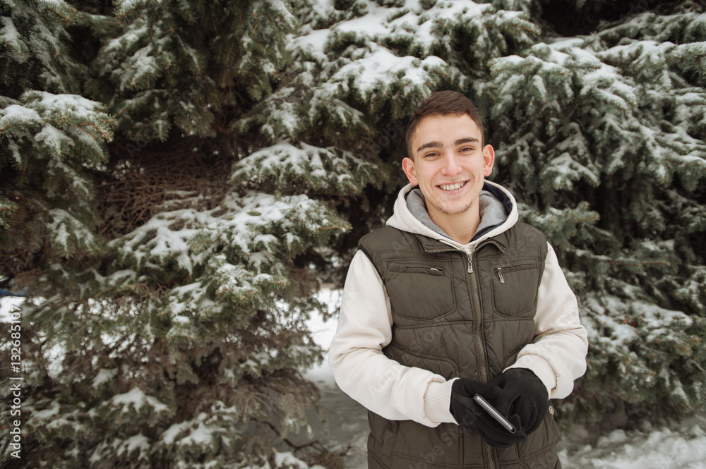 Outdoor winter portrait for young handsome man. Beautiful teenager in his jacket and vest posing on a city street, background of fir trees. Outdoor winter portrait for young handsome man. Beautiful teenager in his jacket and vest posing on a city street, background of fir trees.