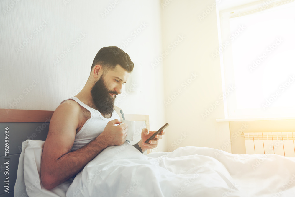 Bearded man lying in bed with morning coffee and phone using app Bearded man lying in bed with morning coffee and phone using app