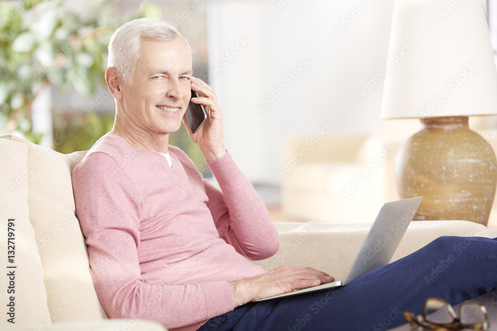 © gzorgz - Telephone banking. Shot of an elderly man sitting with his mobile phone and laptop and banking online.