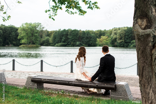 happy bride and groom at a park on their wedding day sitting on a bench near the lake