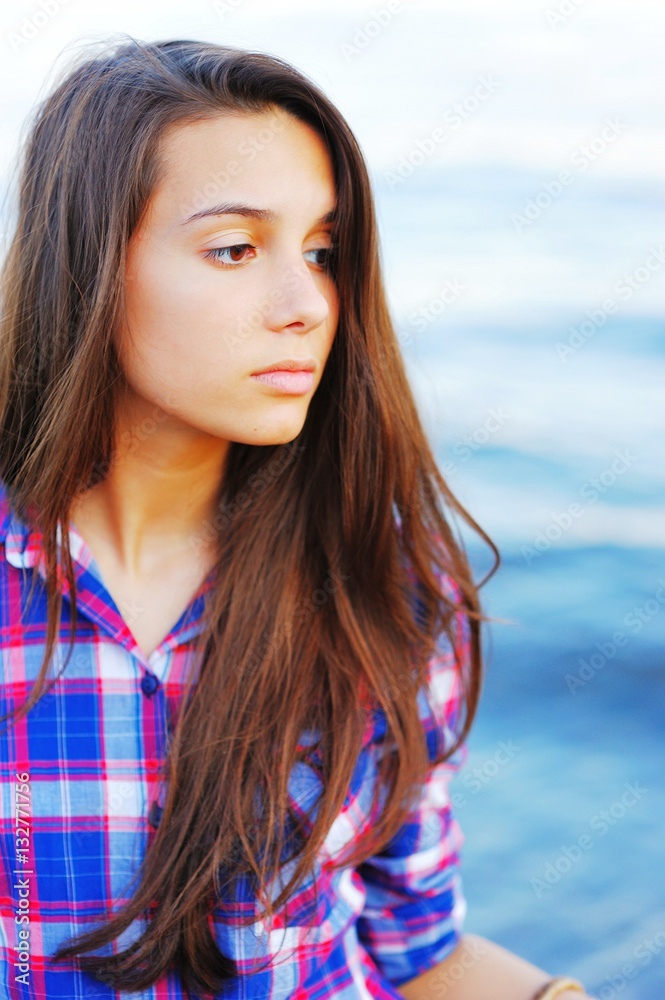 Young pretty brunette girl in red-and-blue plaid shirt with scattered over her shoulders long hair with a thoughtful look on blurred background blue waters of the sea. Young pretty brunette girl in red-and-blue plaid shirt with scattered over her shoulders long hair with a thoughtful look on blurred background blue waters of the sea.