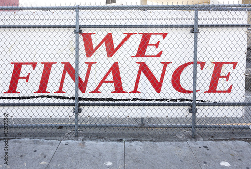 WE FINANCE sign in sketchy urban district behind chain link fence with filthy sidewalk in foreground. Horizontal.