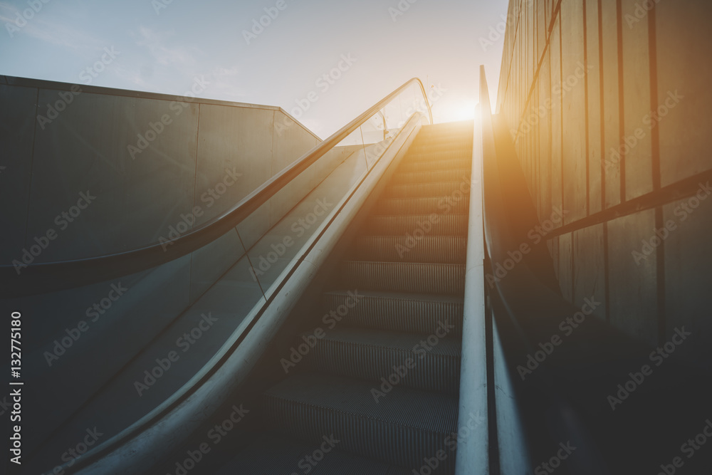 Obraz premium City outdoor grungy modern escalator goes up to sun and teal sky, rusty metal tiled wall on the right, view from the rail, Barcelona, Spain