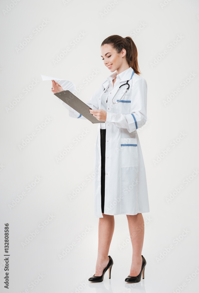 Smiling young woman doctor standing and reading documents on clipboard Smiling young woman doctor standing and reading documents on clipboard