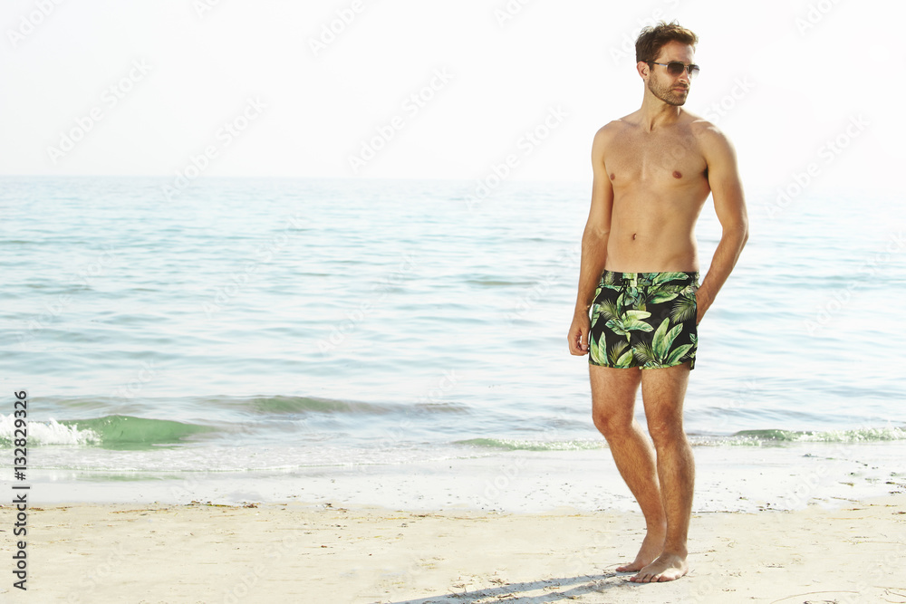 Man shirtless with shorts on beach, looking away
