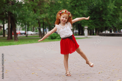 Little smiling girl with red hair poses in green summer park, sh