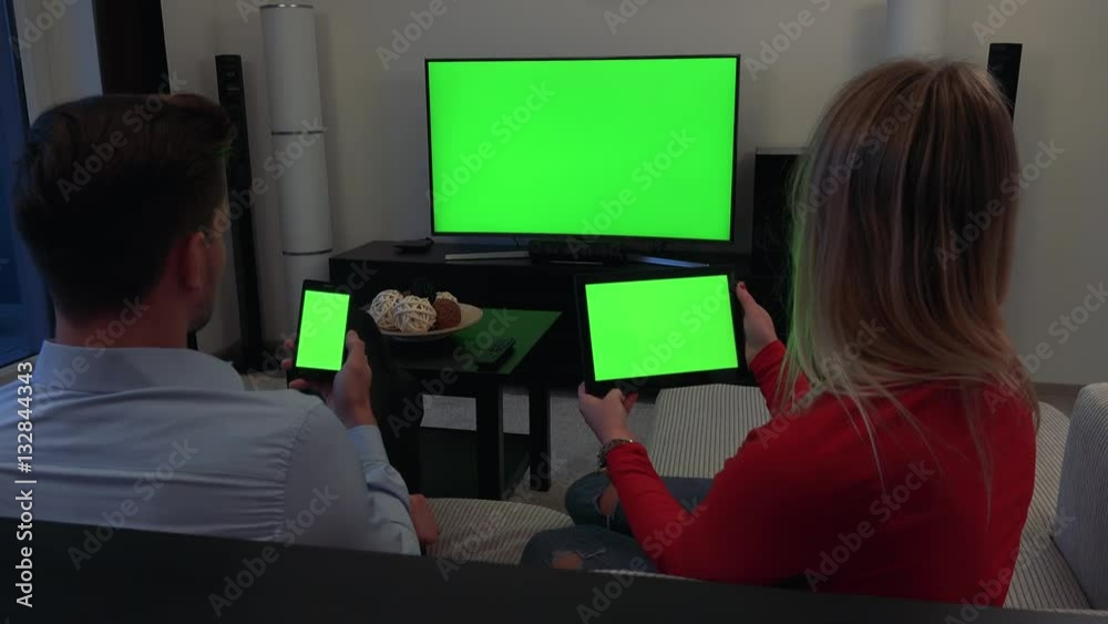 A man and a woman sit on a couch, he holds a smartphone, she holds a tablet, a TV in the background - all three green screens A man and a woman sit on a couch, he holds a smartphone, she holds a tablet, a TV in the background - all three green screens