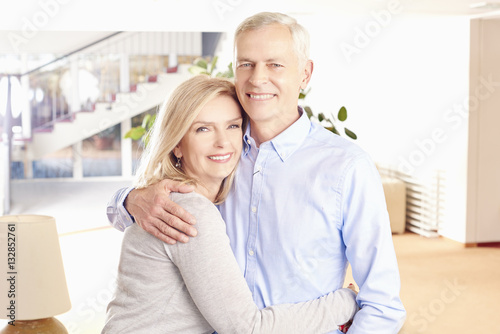 Lovely senior couple. Shot of a happy senior couple standing together. Elderly man embracing her wife from behind while at home in living room.