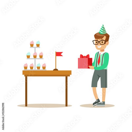 фотографія Boy Holding A Present Standing Next To Table With Cupcakes, Kids Birthday Party