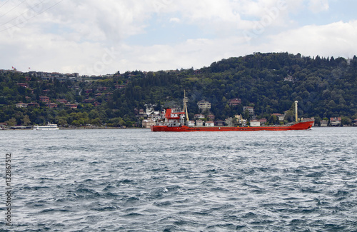 Red, dry cargo ship / vessel crosses Bosphorus strait in Istanbul. Asian side and residential buildings are in the background. It is internationally-significant waterway located in northwestern Turkey