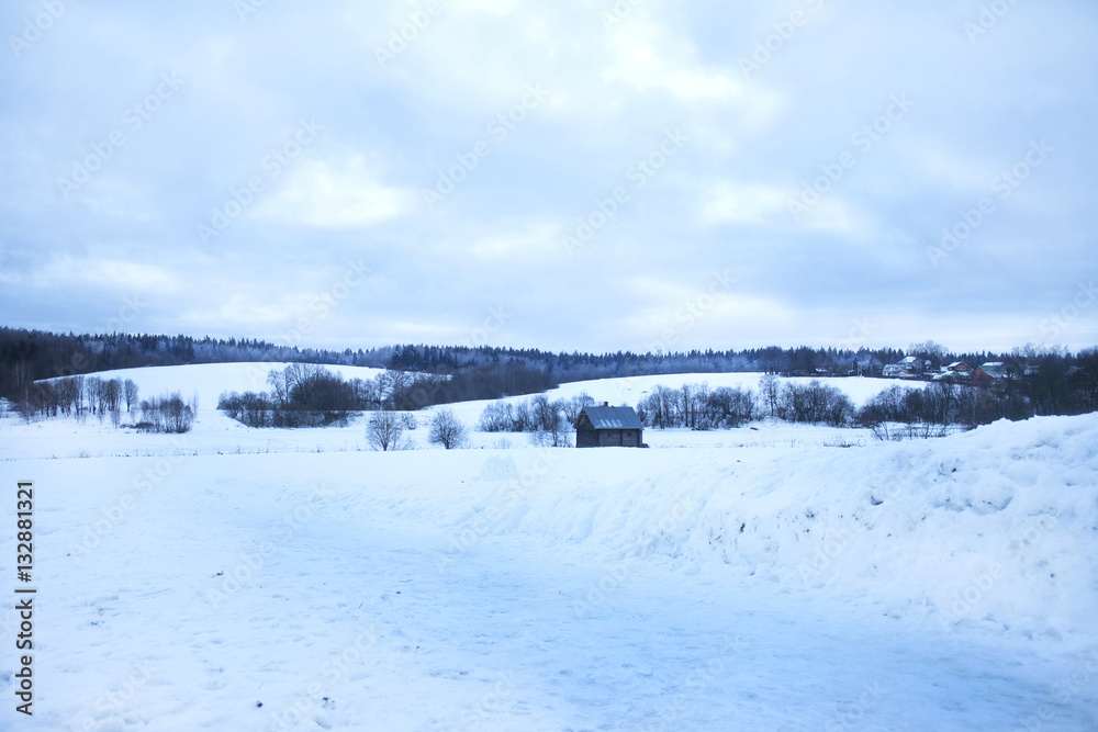 Wooden shed stands in the middle snow, snow-covered hills behind, the forest and the village