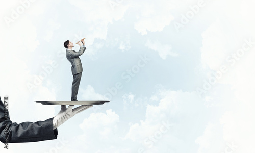 Businessman on metal tray playing fife against blue sky background