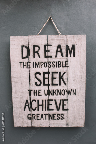Vintage wooden board with motivational phrase hanging on the grey wall. Phrase - Dream the impossible, Seek the unknown, Achieve greatness.