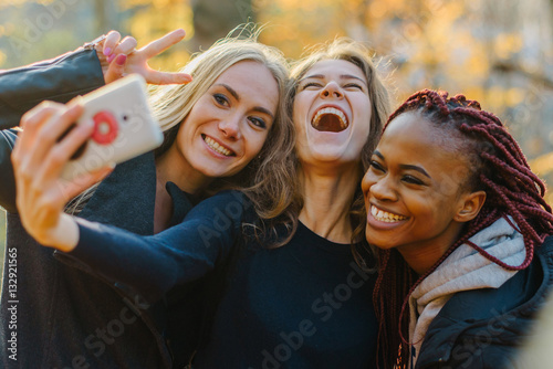 Three Pretty woman making selfie in autumn park. Cute girls with different colored skin. Female making funny faces and smiling at camera photo