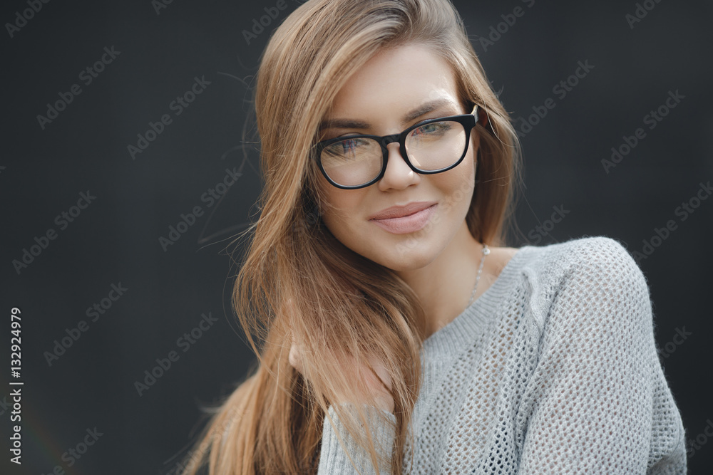 Young beautiful woman with model looks,wears black-rimmed glasses,beautiful makeup,long red hair,big black eye lashes,wearing a light knitted sweater posing in Studio on dark gray background
