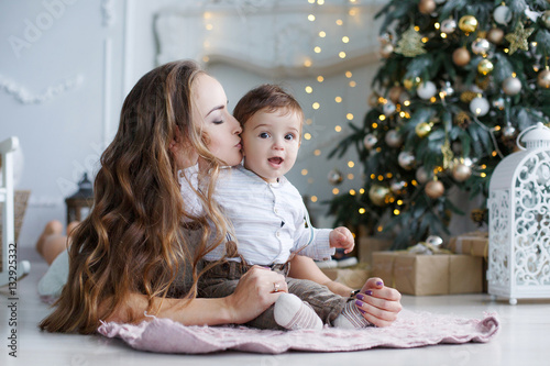 Beautiful woman dressed in a short white lace dress holding New Year s holiday lie on the floor next to a white elegant Christmas tree with his young son Christmas portrait of mother and baby