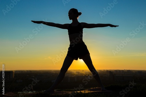 Silhouette of sporty woman doing Ashtanga yoga in the park at sunset. Sunset light, sun lens flares, golden hour. Freedom, health and yoga concept