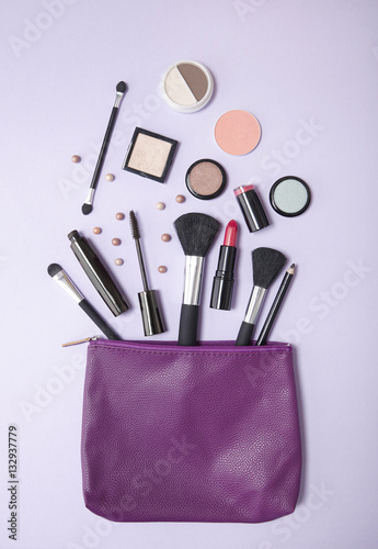 Aerial view of a purple leather make up bag, with cosmetic beauty products spilling out on to a pastel colored background