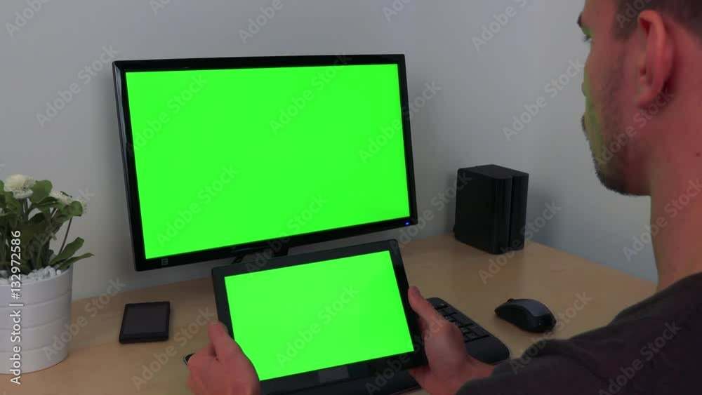 A man watches in turns his tablet and a computer screen, both green screens A man watches in turns his tablet and a computer screen, both green screens
