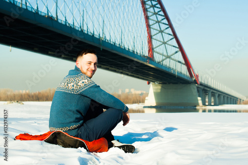man in warm clothes with a beard is walking on the street in the winter in a warm sunny day at the river bridge in the background, sits in the snow