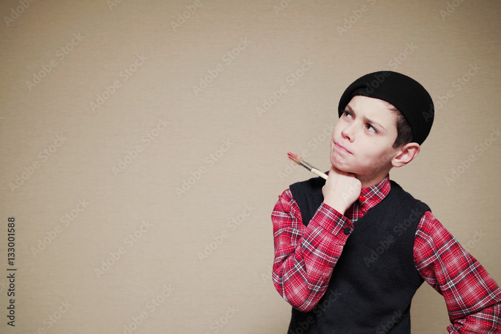 Boy in a beret with a paint brush on a light background. Wondering look. Boy in a beret with a paint brush on a light background. Wondering look.