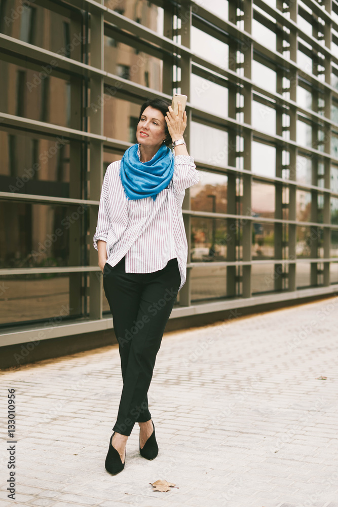 A hipster girl wearing casual clothes is smiling at camera while standing on a street. Young girl is looking aside while walking down the street. Full length portrait of a modern business woman.