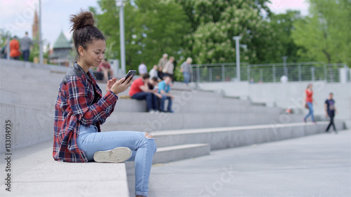 Pretty teenage girl using her mobile phone while sitting outdoors. Casual style - jeans and checkered shirrt.