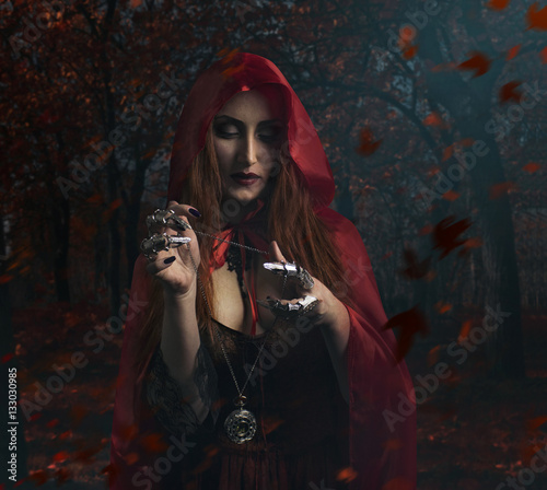 Sorceress in the forest. Beautiful sexy sorceress in red cloak holding antique clock watch standing in the forest photo.