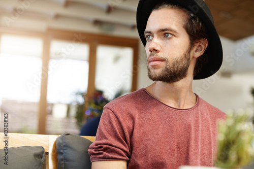 Headshot of handsome young model with fuzzy beard in stylish headwear and t-shirt with rolled up sleeves looking away while posing indoors, sitting on sofa at cafe against blurred interior background