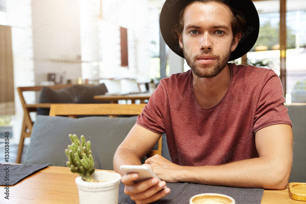 Staying connected. Indoor shot of stylish student in black hat texting friends via social networks, using free wi-fi on his cell phone during breakfast at cafe with modern interior, looking at camera Staying connected. Indoor shot of stylish student in black hat texting friends via social networks, using free wi-fi on his cell phone during breakfast at cafe with modern interior, looking at camera