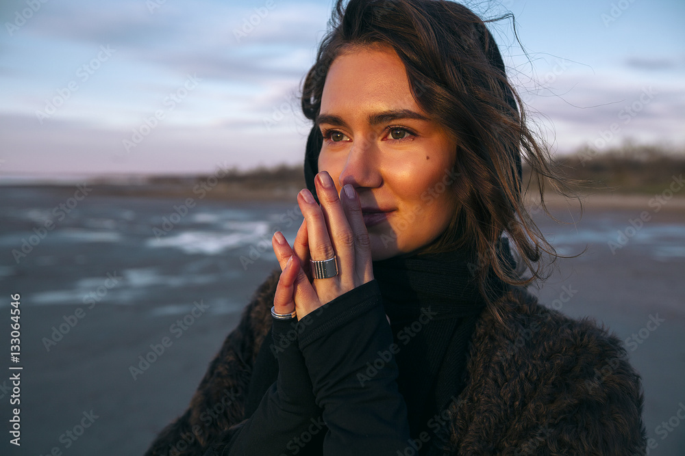 Beautiful girl is standing on the sea in a Sunbeam, yellow boots, long black coat, sand underfoot Beautiful girl is standing on the sea in a Sunbeam, yellow boots, long black coat, sand underfoot