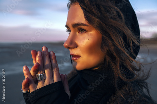 Beautiful girl is standing on the sea in a Sunbeam, yellow boots, long black coat, sand underfoot
