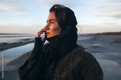Beautiful girl is standing on the sea in a Sunbeam, yellow boots, long black coat, sand underfoot