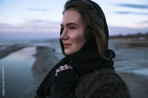 Beautiful girl is standing on the sea in a Sunbeam, yellow boots, long black coat, sand underfoot