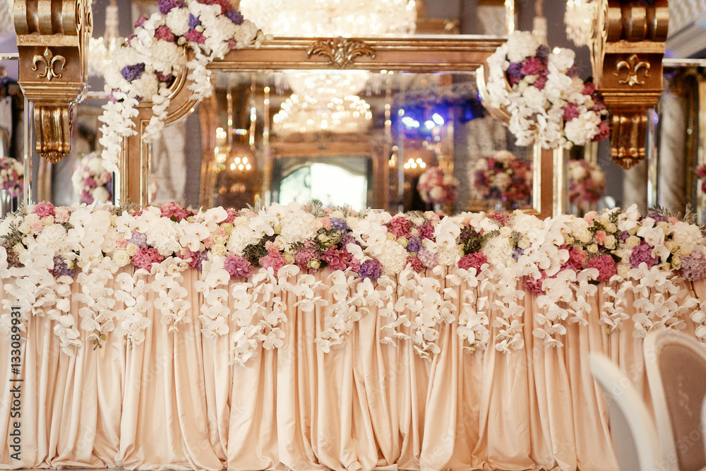 Long dinner table covered with beige cloth and decorated with fl