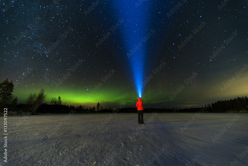 person puts bright blue flashlight to the starry sky with green north lights using torch staying on frozen lake 