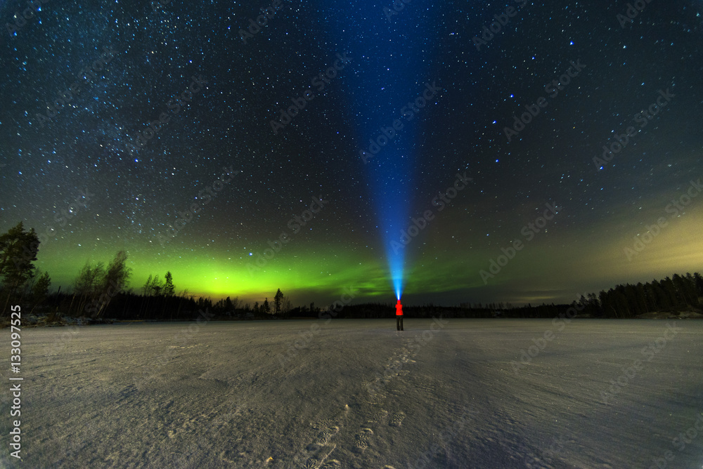 person puts bright blue flashlight to the starry sky with green north lights using torch staying on frozen lake 