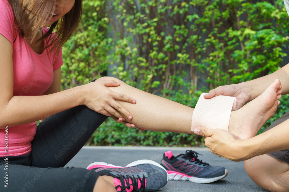 Young woman runner ankle being applied bandage by man in park. 
